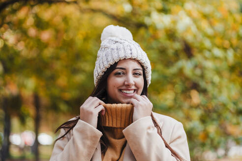 Lächelnde Frau mit Strickmütze hält Pullover im Park - EBBF04823