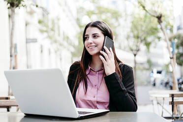 Lächelnde Geschäftsfrau mit Laptop, die in einem Café mit ihrem Mobiltelefon spricht - EBBF04831