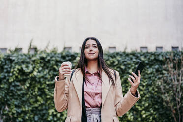 Businesswoman holding disposable coffee cup in front of ivy plants - EBBF04847