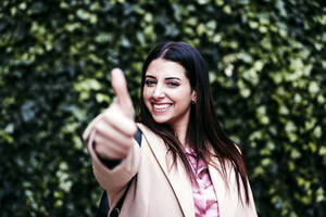 Lächelnde Geschäftsfrau zeigt Daumen hoch vor Efeupflanzen - EBBF04850