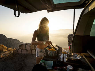 Von hinten beleuchtete Frau, die im Kofferraum eines Lieferwagens bei Sonnenuntergang Essen zubereitet - MALF00374