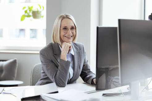 Ältere Geschäftsfrau mit Hand am Kinn am Computer im Büro - FKF04576