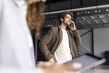 Geschäftsmann im Gespräch mit Smartphone und Kollege mit Tablet-PC im Vordergrund in einer Industriehalle - PESF03269