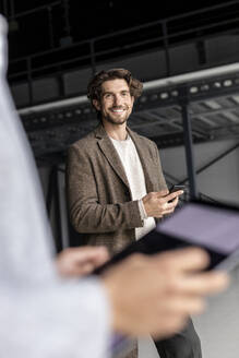 Lächelnder Geschäftsmann mit Smartphone und Geschäftsfrau im Vordergrund in einer Industriehalle - PESF03271