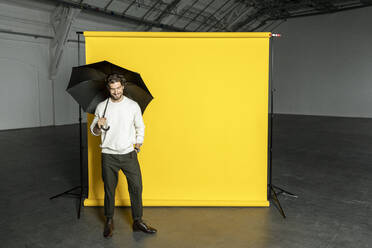 Lächelnder Geschäftsmann mit Regenschirm vor gelbem Hintergrund in einer Industriehalle - PESF03315