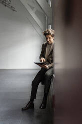 Businessman sitting on window sill and using digital tablet in industrial hall - PESF03335