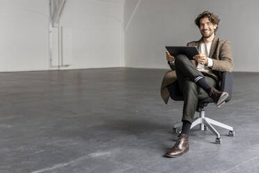 Smiling businessman with digital tablet sitting on chair in empty industrial hall - PESF03337