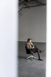 Thoughtful businessman sitting on chair at industrial hall - PESF03339