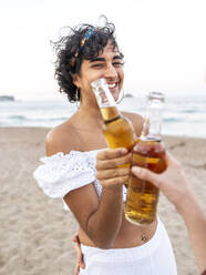 Fröhliche ethnische Frau stößt mit einer Flasche Bier an, während sie einen Sommerabend am Sandstrand genießt - ADSF31705