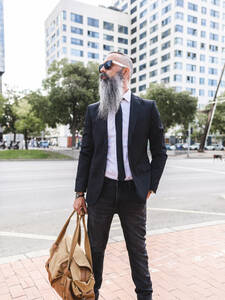 Confident bearded hipster male in classy outfit with bag standing on sidewalk near road on street with buildings in city - ADSF31708