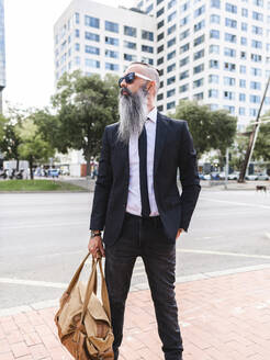 Selbstbewusster bärtiger Hipster in stilvollem Outfit mit Tasche auf dem Bürgersteig in der Nähe der Straße mit Gebäuden in der Stadt stehend - ADSF31708