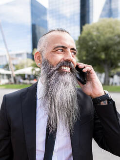 Confident smiling bearded male in classy suit having phone conversation while standing near road on street with modern buildings in city - ADSF31710