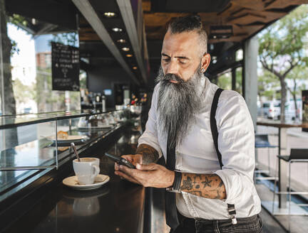 Konzentrierter männlicher Hipster in formeller Kleidung, der mit seinem Handy SMS schreibt, während er mit einer Tasse Kaffee in der Cafeteria am Tresen steht - ADSF31711