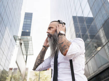 Seitenansicht eines ernsthaften, bärtigen Hipsters in formeller Kleidung, der mit drahtlosen Kopfhörern Musik hört, während er in der Nähe moderner Gebäude steht - ADSF31712