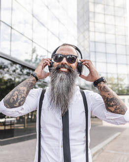 Happy bearded hipster male in formal wear and sunglasses listening to music with wireless headphones while standing near modern buildings - ADSF31713