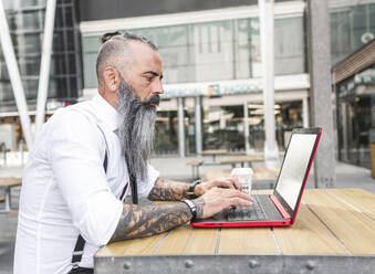 Seitenansicht eines nachdenklichen männlichen Arbeiters in formeller Kleidung, der am Tisch mit Kaffee auf der Terrasse einer Cafeteria sitzt und auf seinem Netbook surft - ADSF31716