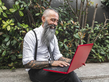 Positiver Hipster männlicher Freiberufler in formeller Kleidung, der ein modernes Netbook durchsucht, während er auf einem Steinrand in der Nähe von Grünpflanzen auf der Straße sitzt, während er online arbeitet - ADSF31718