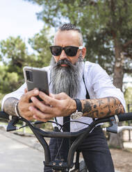 Bärtiger Mann mit Sonnenbrille und weißem Hemd surft auf seinem Smartphone, während er auf einem Fahrrad in einem Park mit Bäumen sitzt - ADSF31721