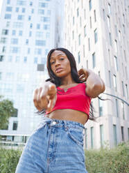 Serious African American female in stylish outfit looking and pointing at camera while standing on street with multistory building on green bushes - ADSF31722
