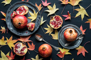 Top view composition of dried autumn leaves placed near plates of ripe sweet pomegranates on black background - ADSF31723