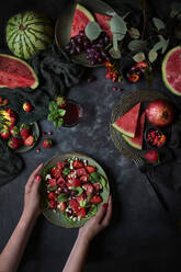 Oben Ansicht der Ernte anonyme weibliche mit Teller mit Wassermelone Salat mit Erdbeeren auf schwarzem Hintergrund mit Granatapfel und Trauben platziert - ADSF31735