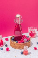 Glasflasche mit frischen Früchten Saft in Holzschale mit reifen Beeren auf dem Tisch mit Gläsern auf rosa Hintergrund serviert - ADSF31744