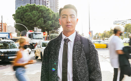 Self assured young Asian male entrepreneur in stylish wear looking at camera against unrecognizable people in sunny city - ADSF31754