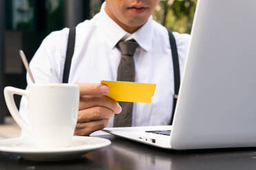 Abgeschnittener, nicht erkennbarer männlicher Unternehmer mit Kreditkarte, der mit einem Netbook und einer Tasse Kaffee am Tisch einer städtischen Cafeteria sitzt und online einkauft - ADSF31756