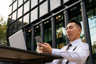 Von unten junger ethnischer männlicher Unternehmer mit Kaffee beim Surfen im Internet auf dem Handy, während er mit seinem Laptop am Tisch einer städtischen Cafeteria sitzt - ADSF31757