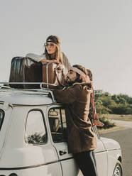 Seitenansicht eines romantischen Hippie-Paares, das auf einem Oldtimer-Auto mit Koffer während eines Ausflugs in die Natur sitzt und wegschaut - ADSF31779