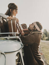 Side view of romantic hippie couple looking at each other while sitting on old timer automobile with suitcase during trip in nature - ADSF31780