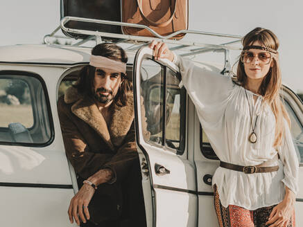 Hippie couple in boho clothes and headbands looking at camera near white old timer automobile during trip together in nature - ADSF31781