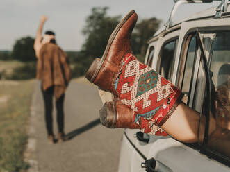 Crop anonymous female hippie in ornamental boots lying in old timer automobile parked on road near faceless man during trip in nature - ADSF31782