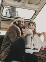 Positive hippie couple in boho styled outfits and headbands sitting in old timer automobile during trip in nature on summer day - ADSF31783