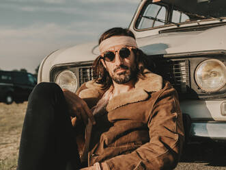 Seriöser männlicher Hippie in Oberbekleidung und Stirnband, der in der Nähe eines weißen Oldtimer-Autos sitzt, während eines Ausflugs auf dem Lande an einem sonnigen Tag - ADSF31784