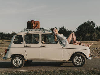 Ganzkörper-Seitenansicht einer barfuß lebenden Hippie-Frau im Boho-Outfit, die sich auf einem weißen Retro-Auto mit Koffer ausruht, während eines Ausflugs in die Natur - ADSF31785