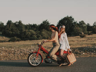 Seitenansicht des Ganzkörpers eines Hippie-Paares, das auf einem roten Moped auf einer asphaltierten Fahrbahn fährt, während eines Ausflugs in die Natur mit Bäumen an einem Sommertag - ADSF31787