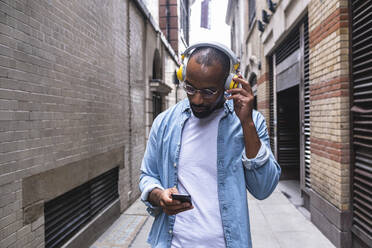 Man with headphones using smart phone walking in alley - ASGF01790