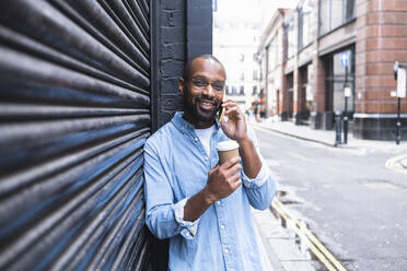 Lächelnder Mann mit Einwegbecher im Gespräch mit Smartphone auf der Straße - ASGF01793