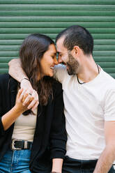 Cheerful enamored young Hispanic couple in casual clothes laughing with closed eyes while hugging and touching foreheads near green wall on city street - ADSF31794