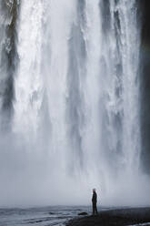 Seitenansicht eines männlichen Wanderers in Freizeitkleidung, der am Flussufer unter dem spektakulären Wasserfall Skogafoss steht, der durch eine massive, mit grünem Moos bewachsene Felswand fließt - ADSF31795