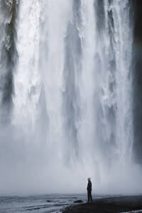 Seitenansicht eines männlichen Wanderers in Freizeitkleidung, der am Flussufer unter dem spektakulären Wasserfall Skogafoss steht, der durch eine massive, mit grünem Moos bewachsene Felswand fließt - ADSF31795