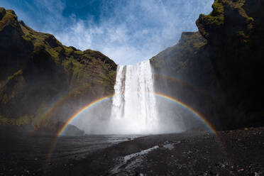 Atemberaubende Szenerie des Regenbogens, der über den reißenden, mächtigen Skogafoss-Wasserfall scheint, der durch die felsige Klippe in Island fließt - ADSF31798