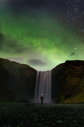 Back view of unrecognizable tourist admiring amazing scenery of powerful Skogafoss waterfall flowing through rocky cliff under magnificent starry sky with polar lights during trip in Iceland - ADSF31799