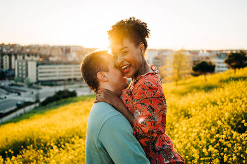 Side view of happy young African American woman with short hair smiling while embracing boyfriend with closed eyes standing in blooming meadow in sunny day - ADSF31801