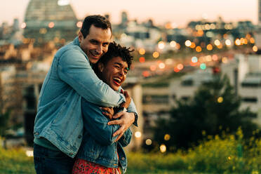 Romantic diverse couple embracing and looking away while standing on lawn against cityscape with buildings on blurred background - ADSF31804