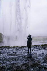 Rückenansicht eines nicht erkennbaren Reisenden in warmer Oberbekleidung und Kapuzenpulli, der den malerischen Seljalandsfoss-Wasserfall während einer Reise in Island mit dem Smartphone fotografiert - ADSF31807