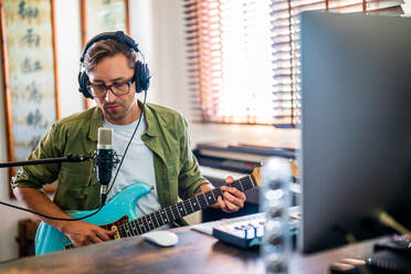 Männlicher Gitarrist mit Brille und Kopfhörern, der ein Instrument hält und einen Computer in einem modernen Studio benutzt - ADSF31810