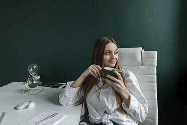 Lächelnde Geschäftsfrau mit Kaffeetasse auf einem Stuhl im Büro sitzend - LLUF00395