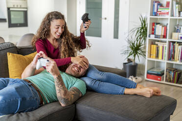 Smiling woman with game controller covering man's eyes on sofa at home - DLTSF02413